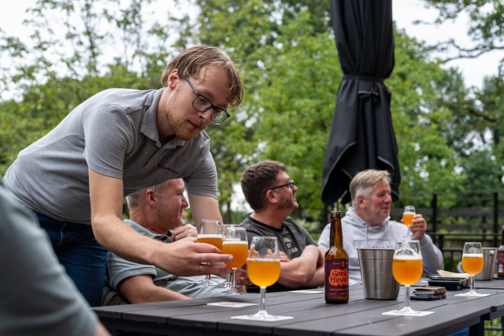 Biersommelier van DeBiermeneer buigt over de tafel en deelt gevulde proefglazen uit aan de groep